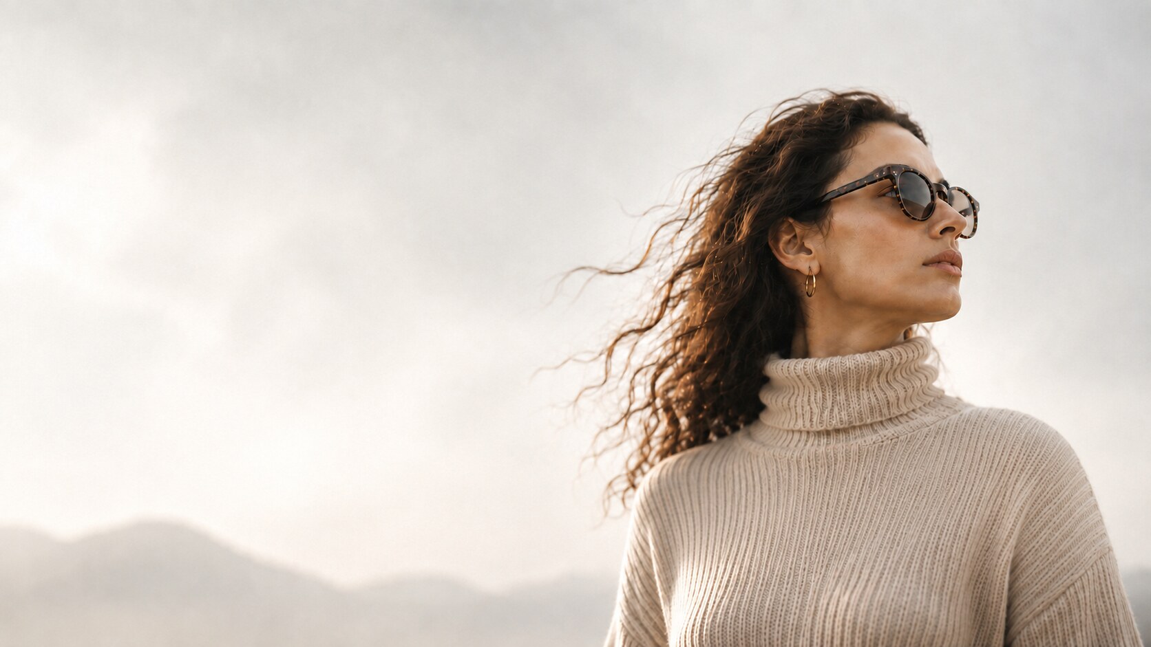 Woman in everyday sunglasses outdoors, hair windblown against a bright hazy sky, illustrating wearable AI that disappears